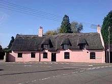 Large pink thatched cottage built on slight curve of road 5 front windows 1 front door 3 attic windows chimney No front garden
