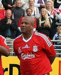 A black man standing with a red shirt. The shirt has emblems in white and the words Carlsberg in the middle.