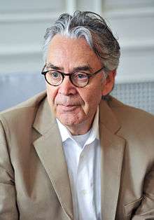 A picture of a man with grey hair. He wears thick-rimmed glasses, a white shirt, and a beige suit.