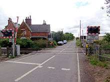 a normal country road passes straight up the middle of the picture, and a two-track railway line crosses it in the foreground.  Modern red-and-white barriers protect the track, and are currently up.  On the left, just beyond the line, 19th century red brick buildings stand next to the track.  This was once the railway station but is now a private house.  The line and the road are fringed with hedgerows, those along the road on the left being particularly tall.  A field of wheat can just be glimpsed on the right, behind the barriers, lights, and hedgerows.