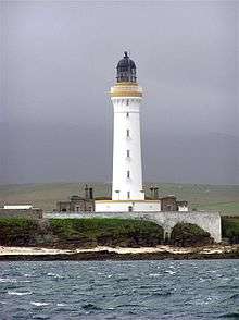  A tall white lighthouse with a brown stripe around the parapet and dark coloured lantern sit on a rocky shore. A white wall obscures the lower floor of grey stone buildings gathered around its base.
