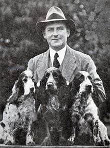 "A greyscale photo of man in a fedora hat and suit with tie standing behind three cocker spaniel with light and dark patches."
