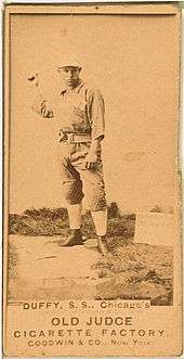 A baseball player is standing in his uniform, with his arm extended in the act of throwing a baseball.