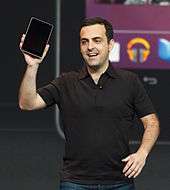 Smiling white man wearing black T-shirt holding a black rectangular device in his right hand.