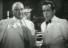 Black-and-white film screenshot of two men, both wearing suits. The man on the left is older and is nearly bald; the man on the right has black hair. In the background several bottles of alcohol can be seen.