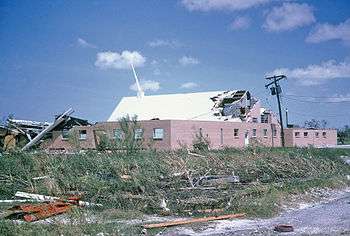 Picture of a building with severe damage to its roof, and debris strewn about in the foreground.