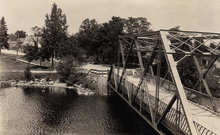 "A sepia monochrome image of a one-lane girder truss bridge over a river. On the opposite shore is a residential community."