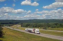 A divided expressway curves from the right of the image through a mostly wooded landscape towards a ridgeline at the rear below a blue sky filled with little white fluffy clouds. Along the roadway closest to the camera is a large white truck with "Perry's Ice Cream" along the side and pictures of scoops of ice cream in various colors and flavors