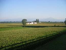 Photo of a rural scene with a farmhouse and crops in the foreground and mountains in the distance.