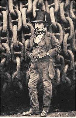 A 19th-century man wearing jacket trousers and waistcoat, hands in pockets, cigar in mouth, wearing a tall stovepipe top hat, standing in front of giant iron chains on a drum.