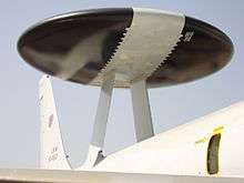 Close-up view of black disc-shaped radar with wide diagonal white band. The radar rests on two convergent struts above aircraft fuselage.
