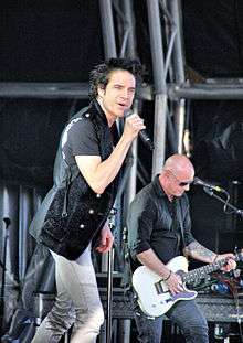A Caucasian man wearing a black jacket sings into a microphone while a bald-headed Caucasian man plays electric guitar.