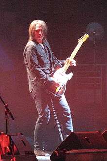 Haug is playing his guitar, while leaning forward and to his right. His hair is shoulder-length, he wears a dark shirt and dark pants. Some band and stage equipment is near his feet.