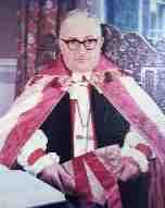 A poor-quality photograph of an old white man seated, wearing spectacles with thick black frames, robed in the vestments of an Anglican bishops: rochet and chimere, pectoral cross, and red-and-white cope; and holding a red-and-gold mitre