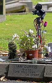A greyish stone block with "Ian Curtis 18-5-80 Love Will Tear Us Apart" carved into it in a sans-serif typeface. There are several small pots of flowers and other objects on top.