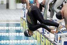 Tall man in black lycra bodysuit with three gold stripes, black cap and goggles, overbalances and falls into the water of a swimming pool off a concrete block. Several other men in bodysuits are standing adjacent to him.