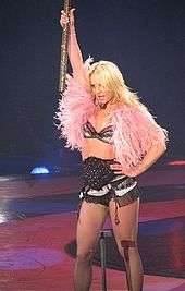 A female blond performer. She wears a black lingerie ensemble underneath a pink fur vest. Her left hand is on her left hip while her right hand is holding a big mallet.