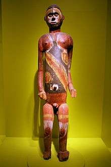 An image of a brown wooden standing male figure partially painted with large black, yellow and white pigment, figure is in an exhibition case on a green background