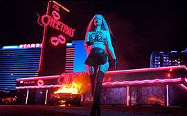 A blond woman in a crop top and mini skirt walking away from a burning car in a parking lot of a bar.