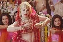 A portrait of a young blonde woman in a red sari and bindi dancing at the forefront of three Indian women performing the same dance.
