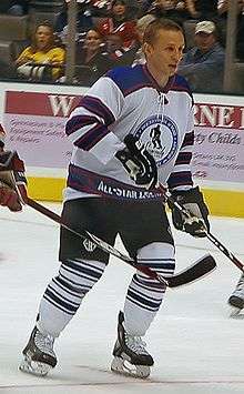 A hockey player with no helmet is skating while looking off into the distance. He wears a white jersey with blue shoulders and three horizontal blue stripes on the sleeves. The logo on his chest is of a silhouetted hockey player skating with a large blue circle around it.