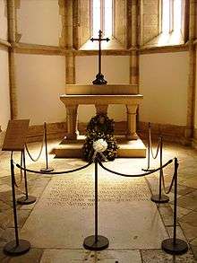 Photograph of a small chapel in which a roped-off inscribed stone slab is set into the floor and with a stone altar in the background on which are a wreath and a large metal cross