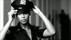 Beyoncé acting like a police officer. She wears the uniform of the New York Police, and she is putting the cap. On the background, a striped wall is visible.