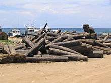 An unorganized pile of approximately 50 rosewood logs sits on a beach with boats in the background.