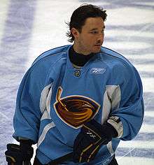A Caucasian hockey player shown from the waist up. He is helmet-less and wears a blue jersey with a stylized brown thrasher holding a hockey stick as a logo