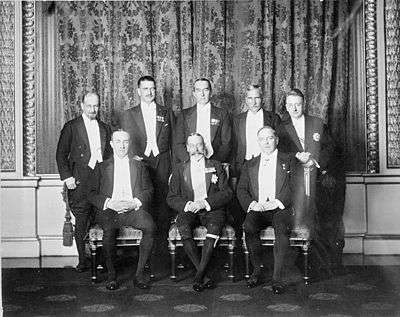 A group pose of eight men in smart evening wear. The King sits in the middle surrounded by his prime ministers.
