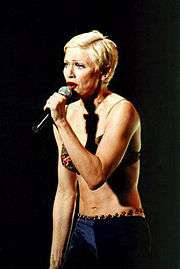 Image of a blond female facing her left. She has short blonde hair and is wearing a green bra and purple pants with beads on the waist. She's singing to a microphone, which she holds to her mouth with her left arm.