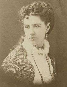 A monochrome photograph portrait of a woman 29 or 30 years old, shown from the chest up, wearing a long necklace with dark beads atop a white blouse with an encircling collar made of lace, covered on the shoulders with a dark lace drape, with long, dark hair curled and secured behind the head with tresses down past the shoulder blades, the woman's body turned to the right but her head turned to the left to reveal a dangling earring.