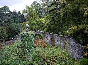 An overgrown area with a circular stone structure covered in ivy in the foreground and a wall with wide embrasures beyond. Tall trees, both conifers and deciduous overlook the scene.