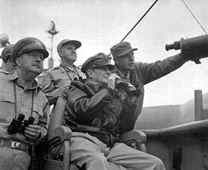 MacArthur wears a bomber jacket and his distinctive cap and holds a pair of binoculars. Almond is pointing something out to him.