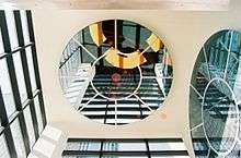 Two circular mirrors with circle and wagon spoke designs are set into an ivory colored ceiling and wall. They reflect windows in black rectangular frames in two other walls.