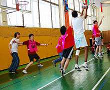 High school or university aged men playing netball on an indoor court. Floor is painted wood. Gym has a window behind the basket.