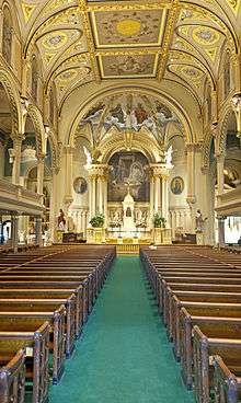A green-carpeted aisle between two sets of wooden pews with a golden-colored decorated ceiling, arches over the side galleries and an arched, painted nave at the rear