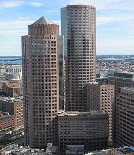 Aerial view of two skyscrapers; both have circular footprints and light red facades. The shorter of the two buildings, in the foreground, is topped by a pyramid-like cap, while the taller building has a flat roof.