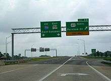 Two green signs are located above an elevated portion of roadway with no traffic visible on a cloudy day.