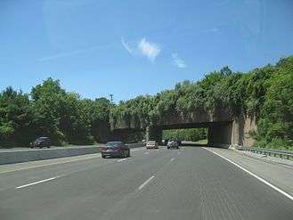 A six lane freeway in a wooded area with an overpass containing trees