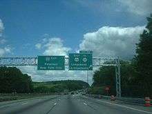 A six lane freeway in a wooded area approaching an interchange with two green signs over the road. The left sign reads Interstate 80 east Paterson New York City and the right sign reads exit 28 east U.S. Route 46 to Route 10 Ledgewood Lake Hopatcong upper right arrow.