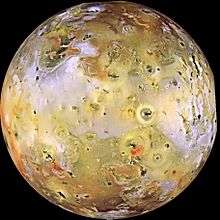 A multi-colored image of the full disk of a planetary body, dotted with numerous dark spots. Much of the middle portion of the planetary body is yellow to white/gray, while the polar regions at the top and bottom are generally reddish in color.