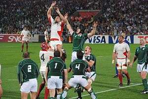 Two rows of opposing players, green to the fore, white behind, each aid a jumping player from their team by lifting him towards an off-picture ball travelling overhead.