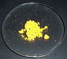Some canary-yellow powder sits, mostly in lumps, on a laboratory watch glass.