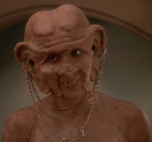 A Caucasoid woman in the heavy orange makeup and protheses typical of Ferengi looks to the camera's left; she is nude, baring her upper chest and shoulders