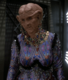 A Caucasoid woman in the heavy orange makeup and protheses typical of Ferengi looks to the camera's left; she is wearing a garish, skintight outfit and excessive jewelry from her clothes, neck, and lobes