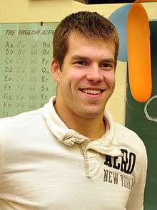 A man standing directly in front of the camera. He has short brown hair and is wearing a light colored polo shirt.