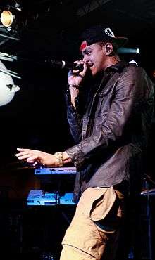 A man wearing a cap and a black leather jacket raps into a microphone.