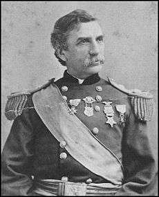 Head and torso of a white man with a mustache and wavy hair wearing an ornate military jacket. The jacket has fringed shoulder boards, a wide sash across the chest, and four medals pinned to the center and left half of the chest.