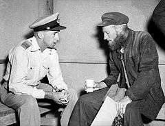 Two seated men in conversation, one clean-shaven and wearing light-coloured military uniform with peaked cap, the other bearded and wearing dark overalls and cap